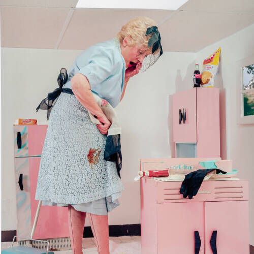 Color photograph of a woman, surprised, at a stain on her dress. She mis much larger than the pink kitchen items around her.
